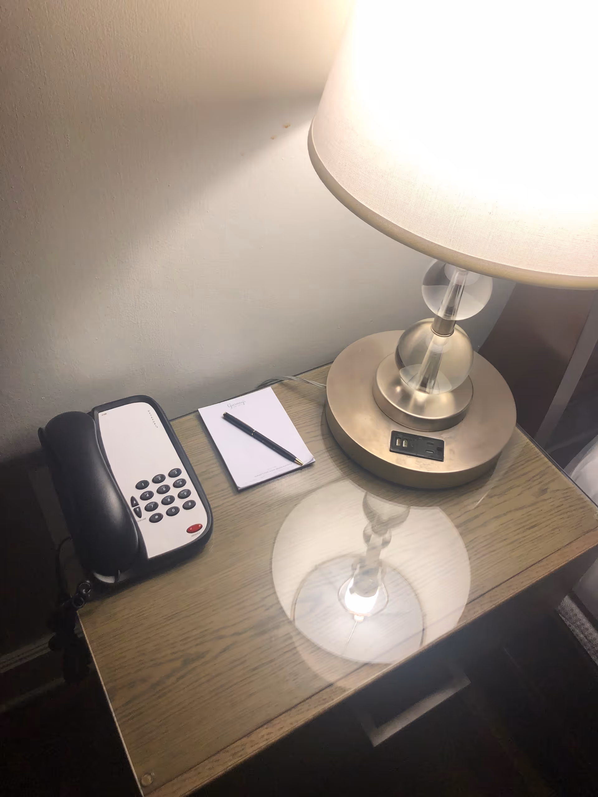 A wooden bedside table with a glass top, holding a lamp with a beige shade and a built-in power outlet, a black and white telephone, and a small notepad with a pen on top.