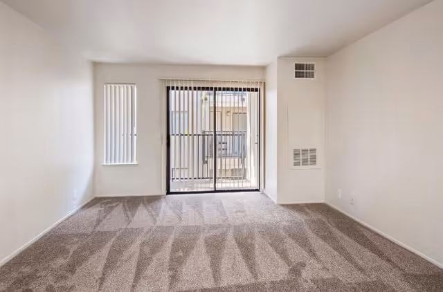 Empty carpeted living room with a sliding glass door and vertical blinds.