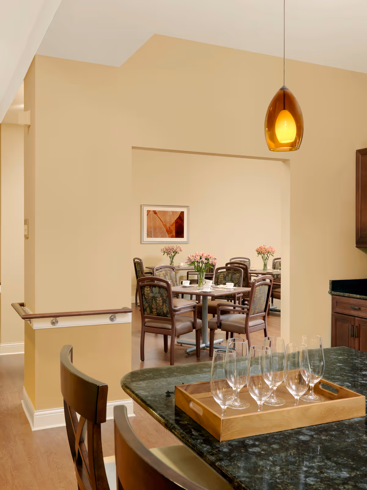 Interior view of a dining area in a senior living facility showing a table with six chairs and flower vases, seen through an open doorway from a kitchen counter with a wooden tray holding six empty wine glasses. The walls are painted beige, and there is a hanging amber pendant light above the counter.