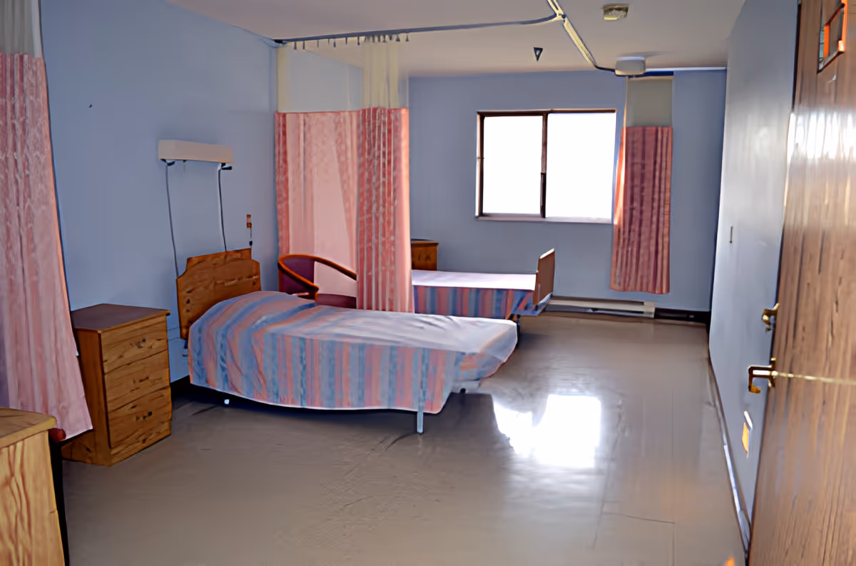 A sparse shared bedroom with two single beds, wooden dressers, pink curtains, and a window at the far wall.