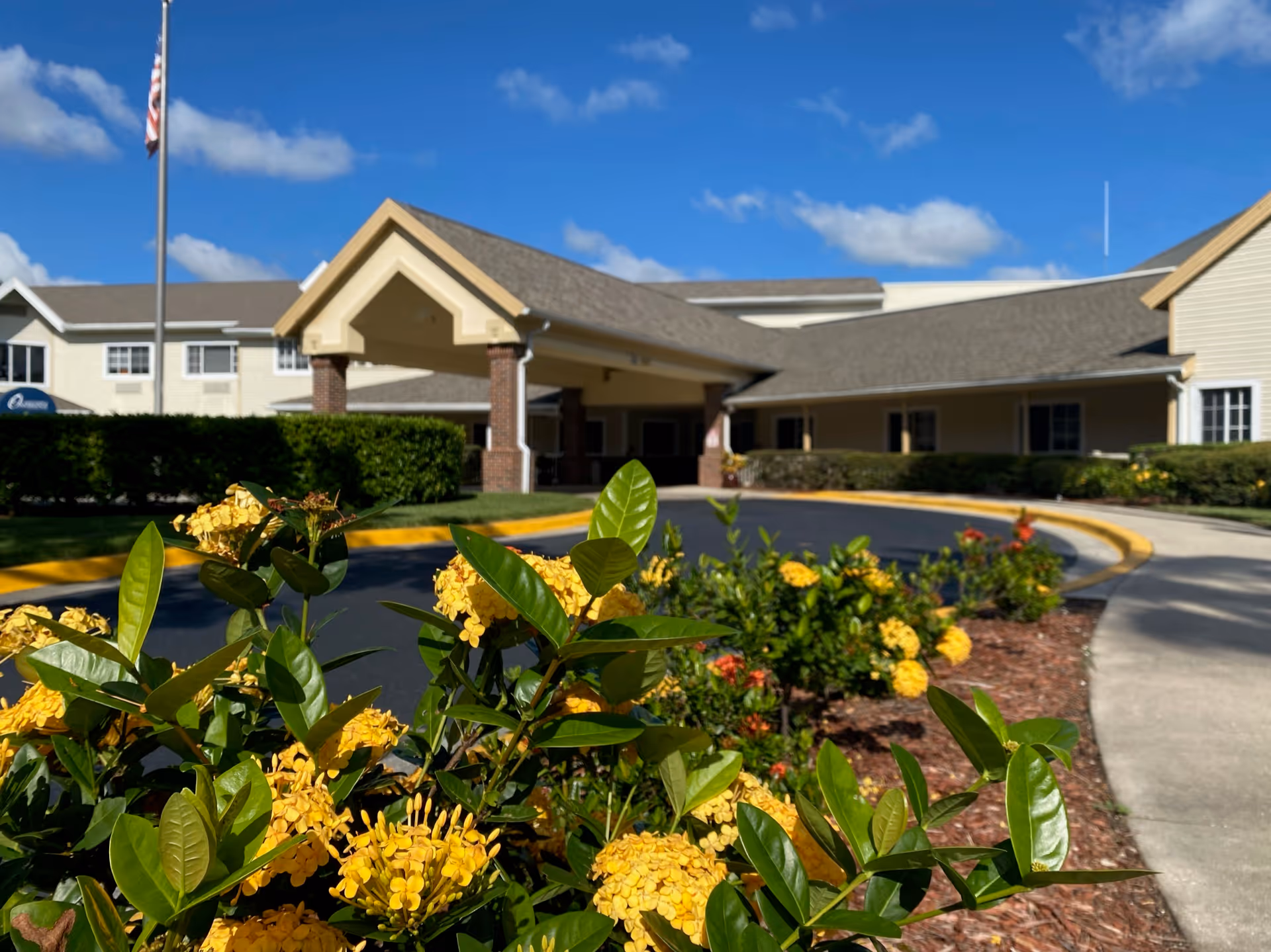 Front entrance of a senior living building with a covered drop-off and yellow flowering shrubs in the foreground.