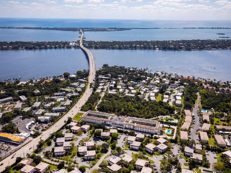 Aerial view of a coastal residential area with a long bridge extending over the water connecting two land masses. The image shows numerous buildings, roads, and green spaces, with water bodies on both sides under a partly cloudy sky.