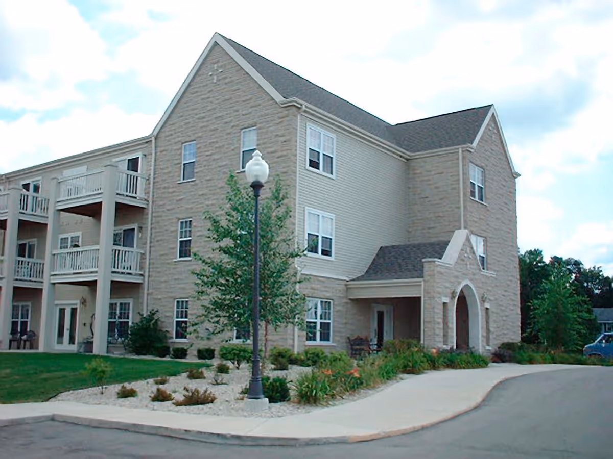 Exterior view of a three-story senior living apartment building with beige stone and siding facade, balconies, a lamp post, landscaped garden beds, and a curved driveway.