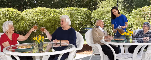 Elderly residents dine outdoors at round tables with a staff member serving.