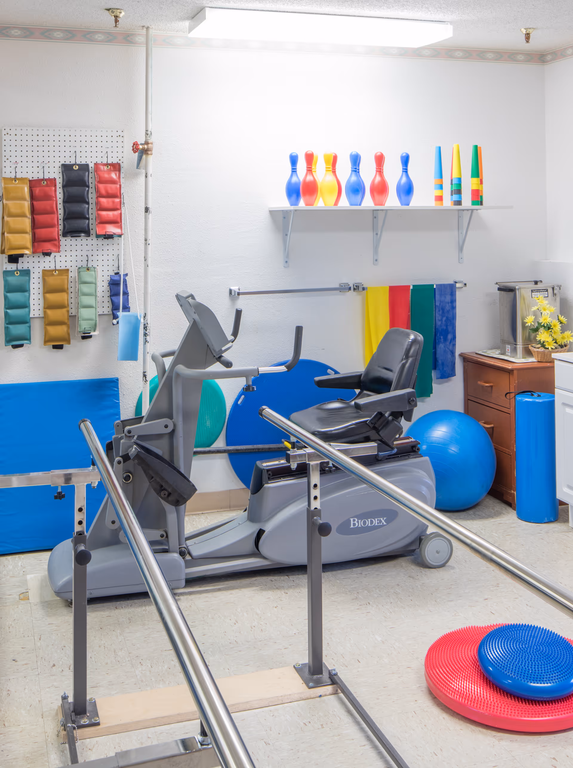 A physical therapy room with exercise equipment including a Biodex recumbent bike, parallel bars for walking support, colorful therapy balls, balance cushions, and pegboards with weighted bags hanging on the wall. There are also colorful plastic bowling pins on a shelf and a wooden cabinet with a water dispenser and flowers.