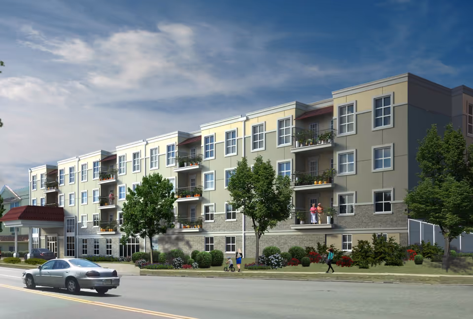Exterior view of a multi-story residential building with balconies, trees, and landscaping along the sidewalk. Several people are visible walking or standing near the building. A car is driving on the road in front of the building under a partly cloudy sky.