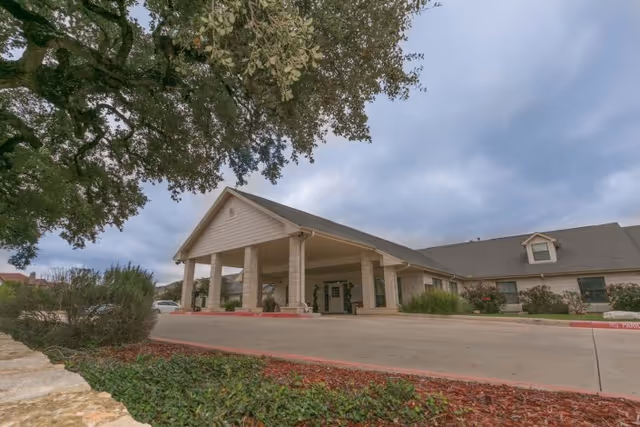 Covered front entrance and porte-cochere of a single-story health center with a driveway, trees, and landscaping under a cloudy sky.