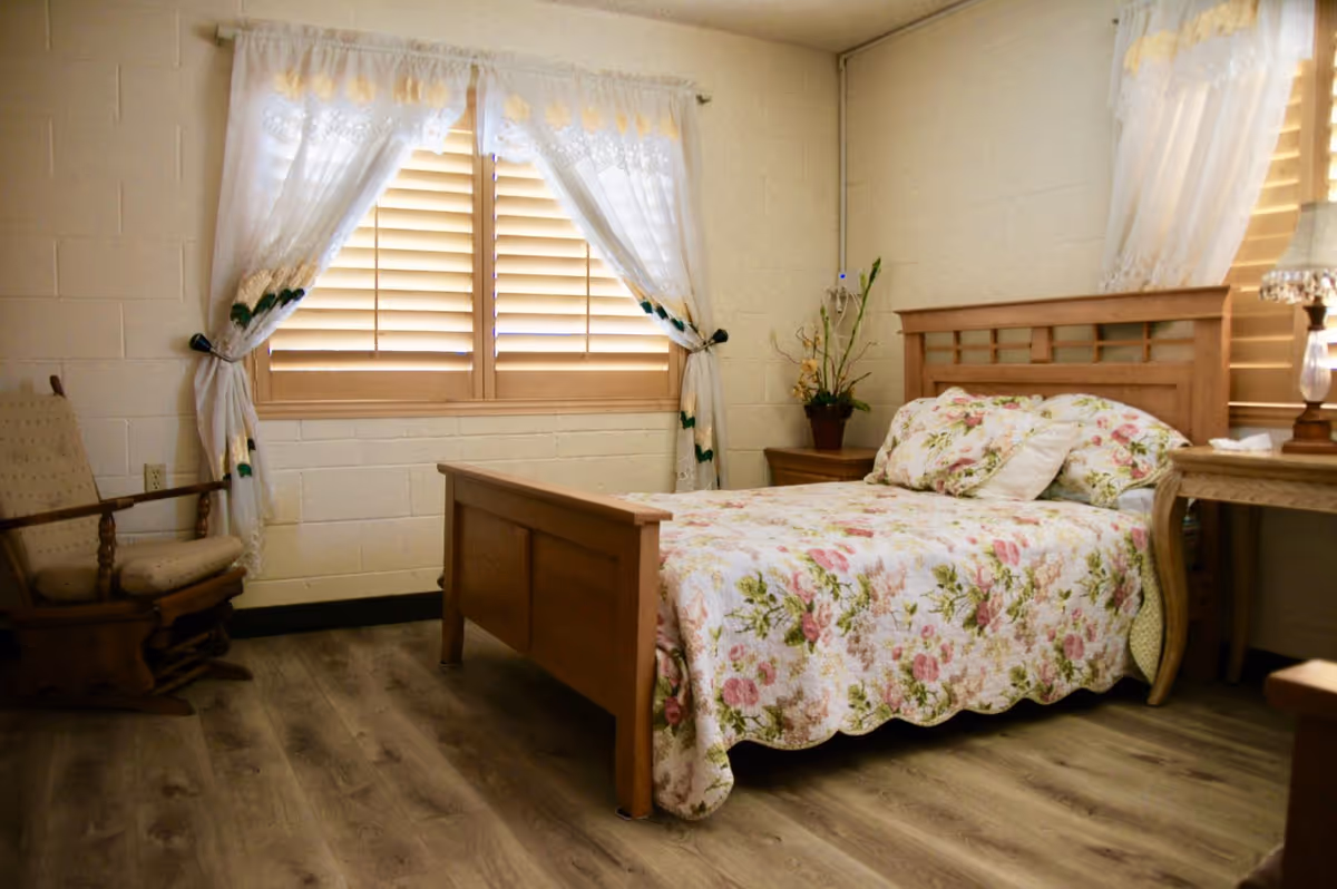Bedroom in a senior facility featuring a floral quilt, wooden bed, and curtained window.
