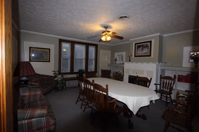 A cozy common room with a long rectangular table covered with a white tablecloth surrounded by wooden chairs. The room has a ceiling fan with lights, three large windows with wooden frames, a plaid sofa and armchair, a lamp with a red shade, plants, and built-in shelves with books and decorative items. The walls are painted light gray with white trim, and there is a framed painting above a white fireplace mantel.