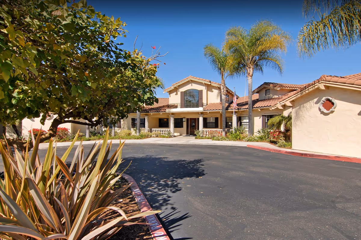 Sunlit exterior of a memory care facility with tiled roof, palm trees, and driveway