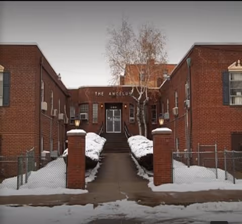 Snow-covered sidewalk and steps lead between brick pillars and chain-link fences to the front entrance of a two-story red brick building with signage above the door.