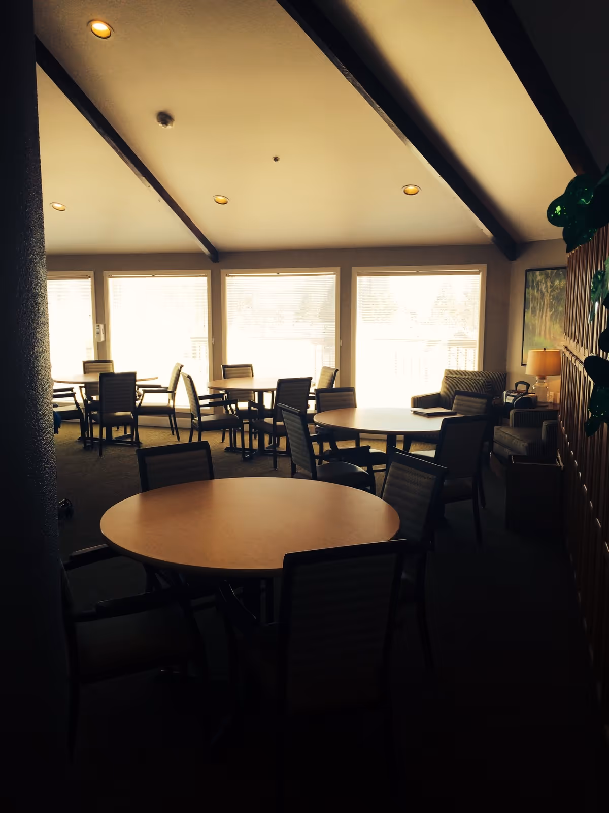 Interior view of a common area with multiple round tables and chairs arranged for seating. The room has large windows letting in natural light, a high ceiling with exposed beams, and a cozy seating area with armchairs and a lamp in the corner.