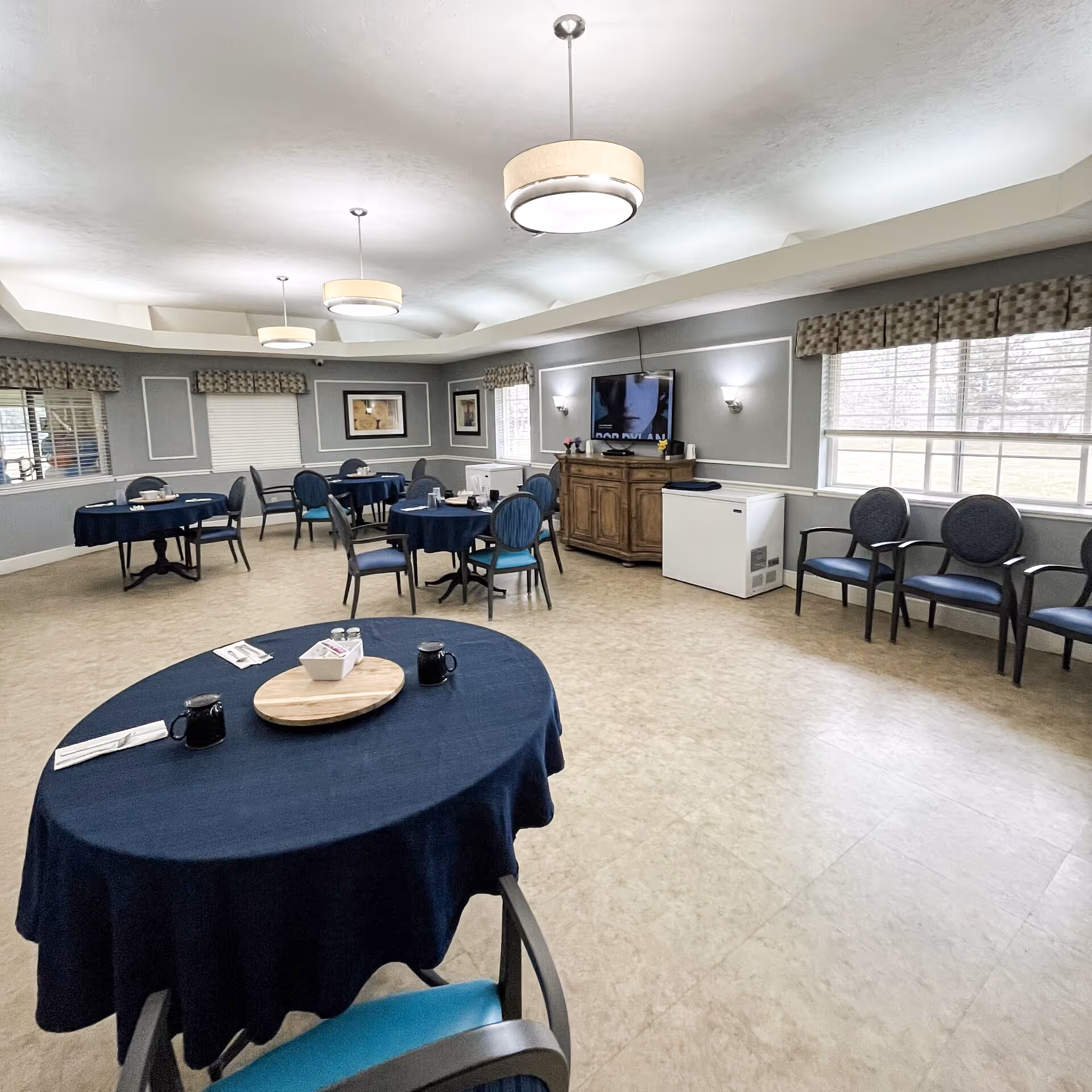 A senior living facility dining room with several round tables covered in dark blue tablecloths, each set with mugs, napkins, and condiments. The room has gray walls with white trim, multiple windows with patterned valances, and three modern ceiling lights. There is a wooden cabinet with a TV mounted above it and a white freezer next to it. Several blue cushioned chairs are arranged around the tables and along the wall.