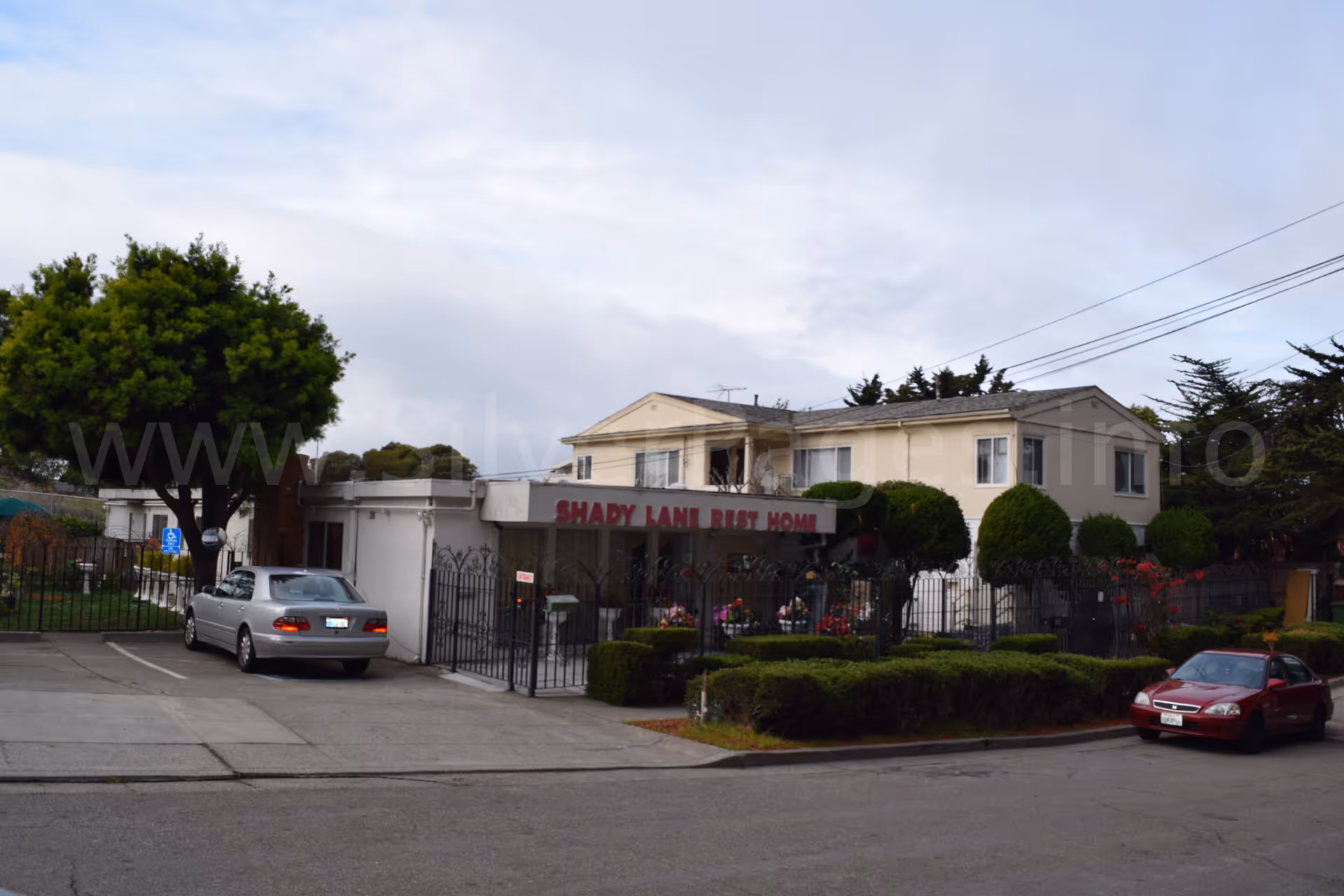 Front exterior of Shady Lane Rest Home with gate, shrubs, and parked cars.