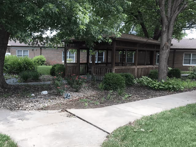 A wooden gazebo surrounded by green bushes, plants, and trees in an outdoor garden area next to a brick building. A concrete pathway curves in front of the gazebo.
