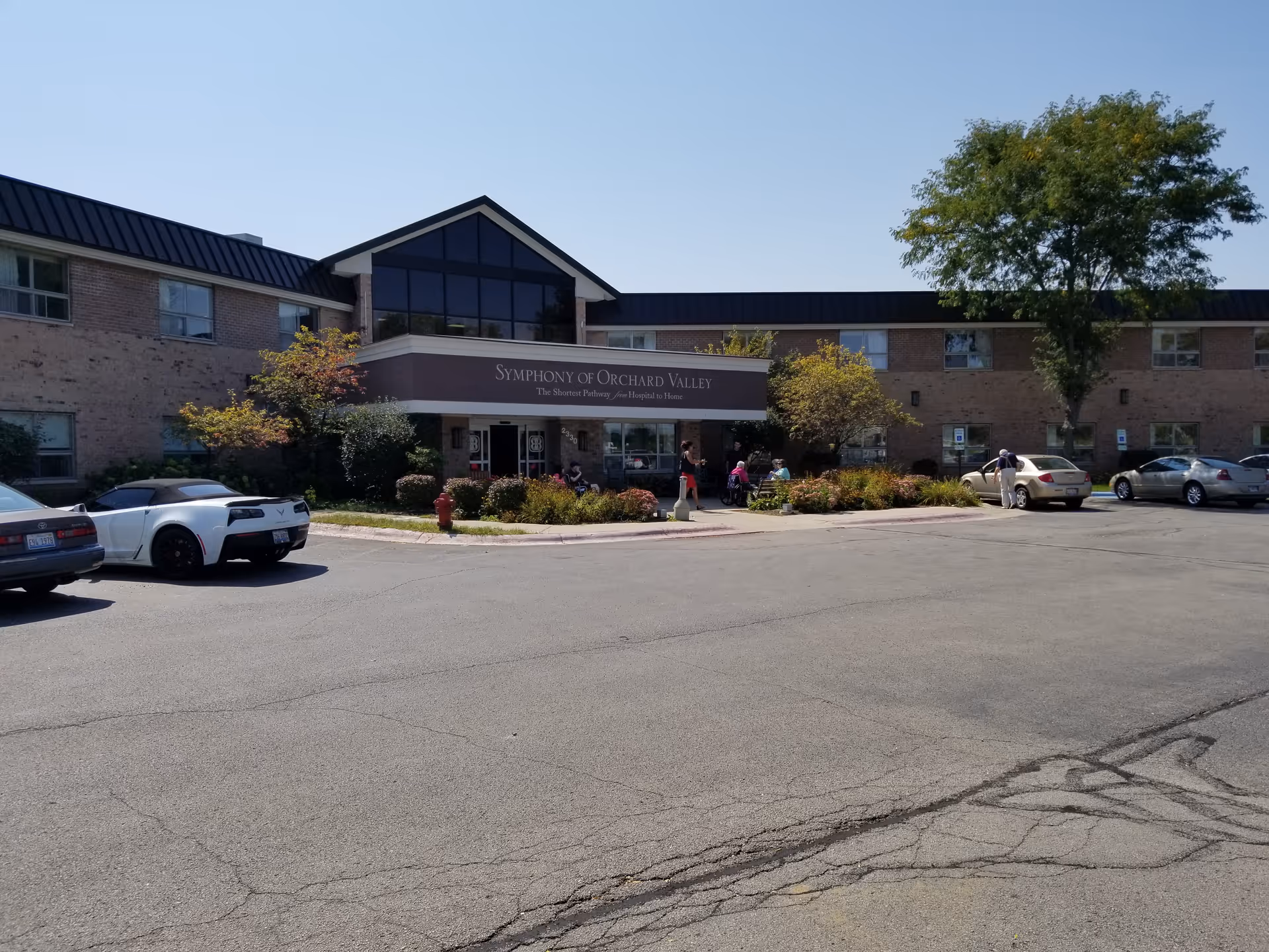 Exterior view of Symphony of Orchard Valley senior living facility with entrance and parked cars.