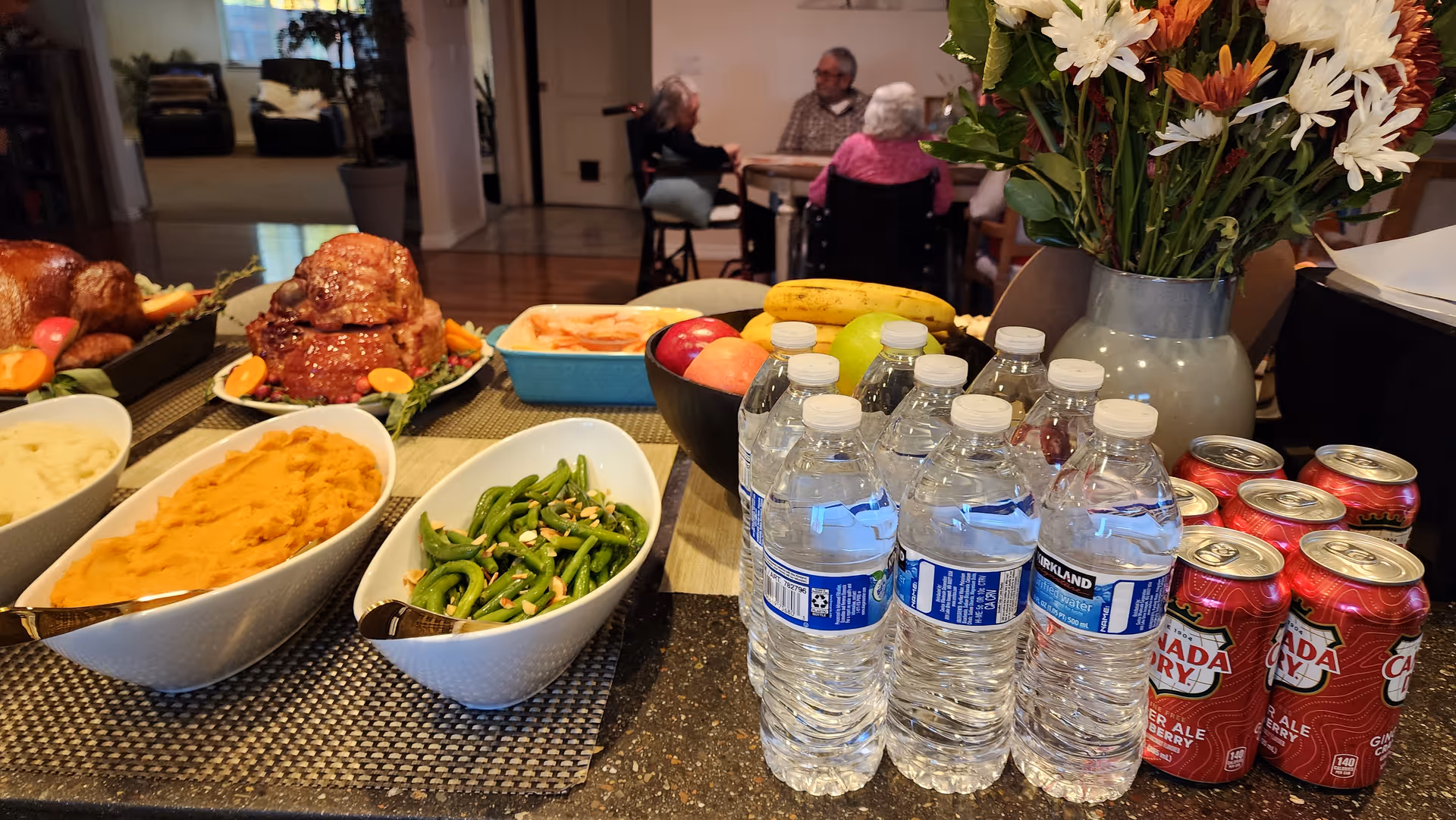 Buffet-style dining table with roasted chicken, vegetables, water bottles, and flowers; residents dining in background.