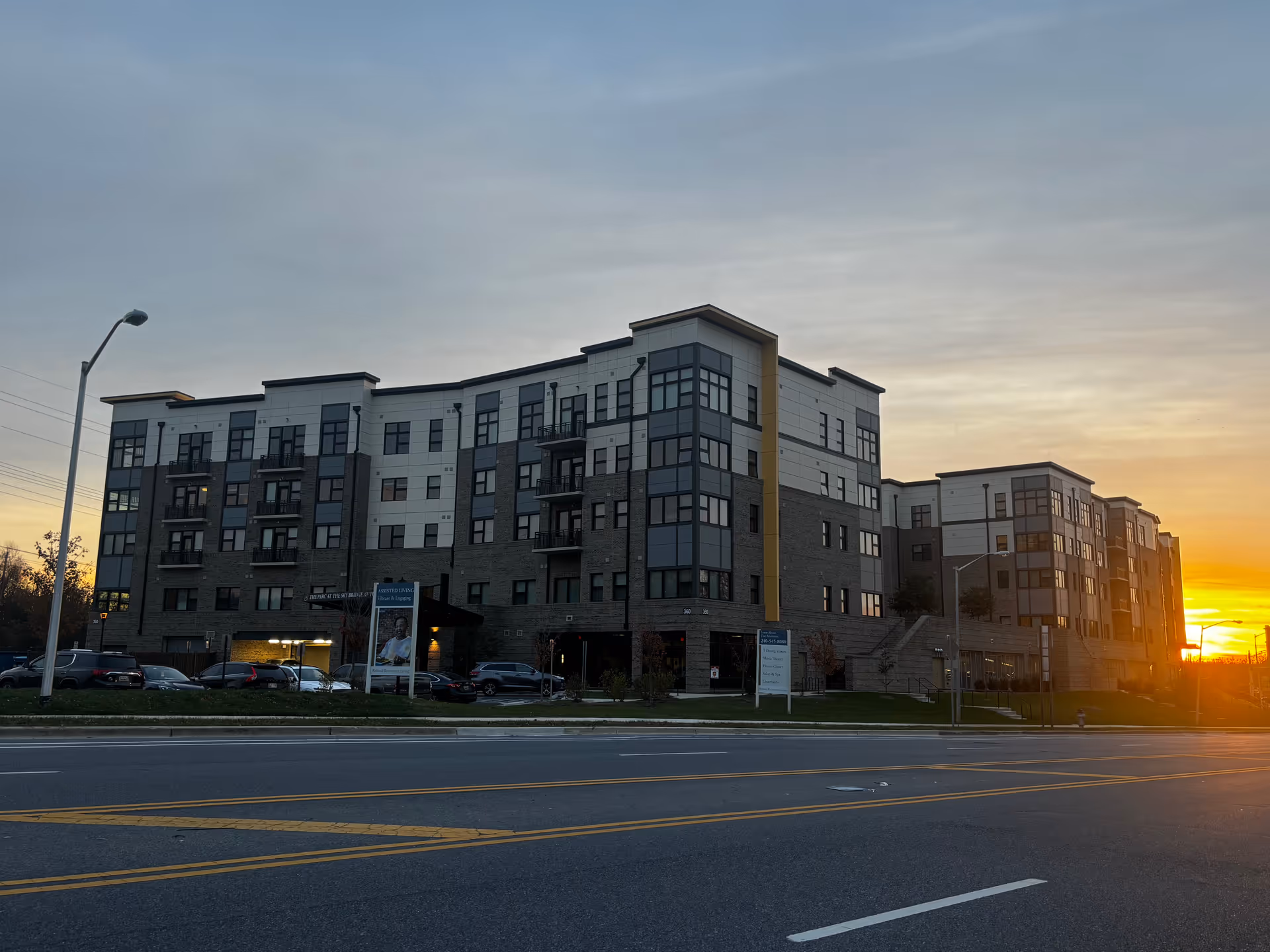 Exterior view of a multi-story senior living building at sunset with parked cars and a street in front.