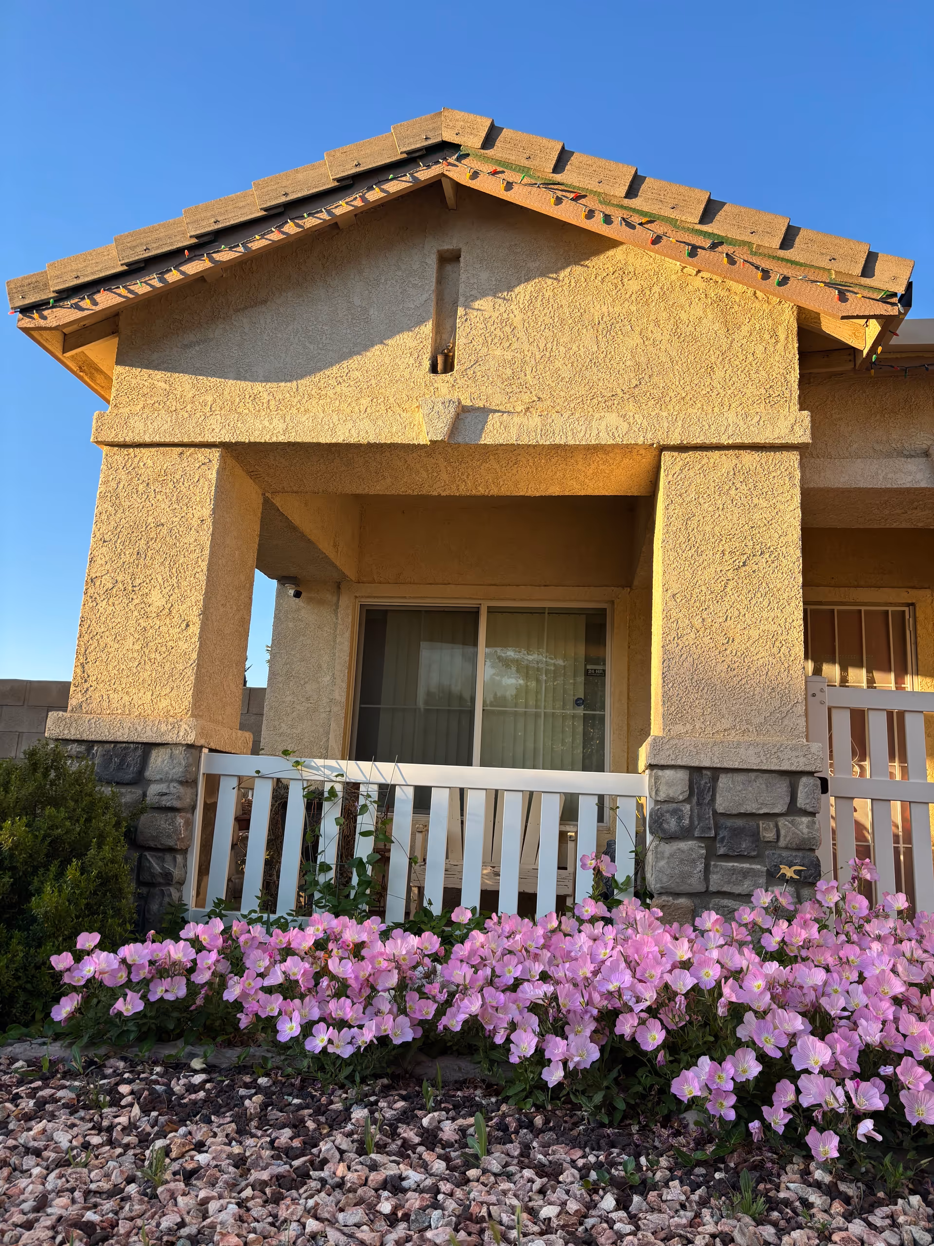 Beige stucco senior living entrance with stone columns, front porch, and pink flowers along the border.