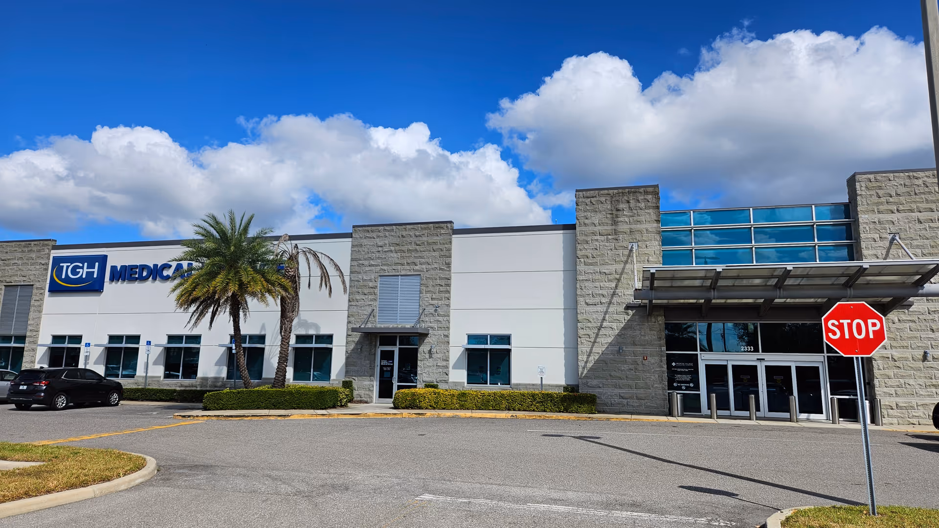 Exterior view of a modern medical facility with palm trees, blue sky, and a STOP sign.