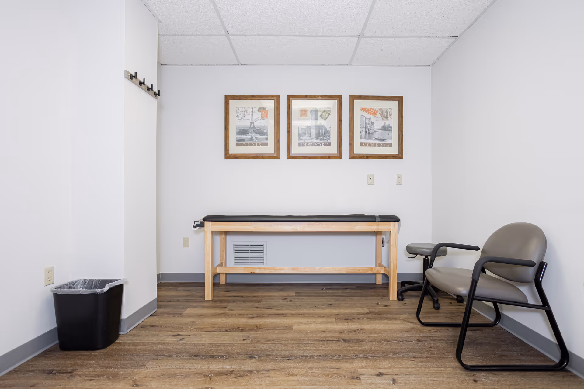 A small medical examination room with a wooden table, chairs, and framed wall art.