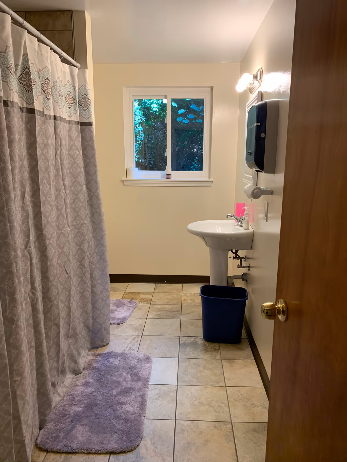A bathroom with shower curtain, pedestal sink, window, purple mats, and a blue trash bin.