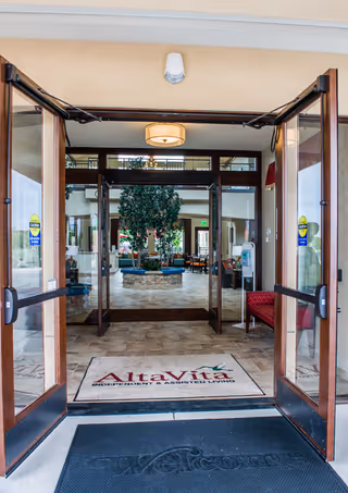Open glass entry doors reveal the AltaVita Assisted Living lobby with a welcome mat, indoor plant and seating area.