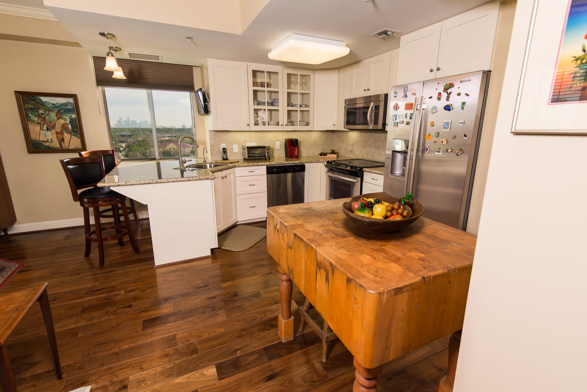 A modern kitchen with white cabinets, stainless steel appliances including a refrigerator covered with magnets, a microwave, and a dishwasher. There is a wooden island table with a bowl of artificial fruit in the center. The kitchen has a granite countertop with a sink and two bar stools. A window with a view of a city skyline is visible in the background, along with a painting on the wall and a mounted TV.