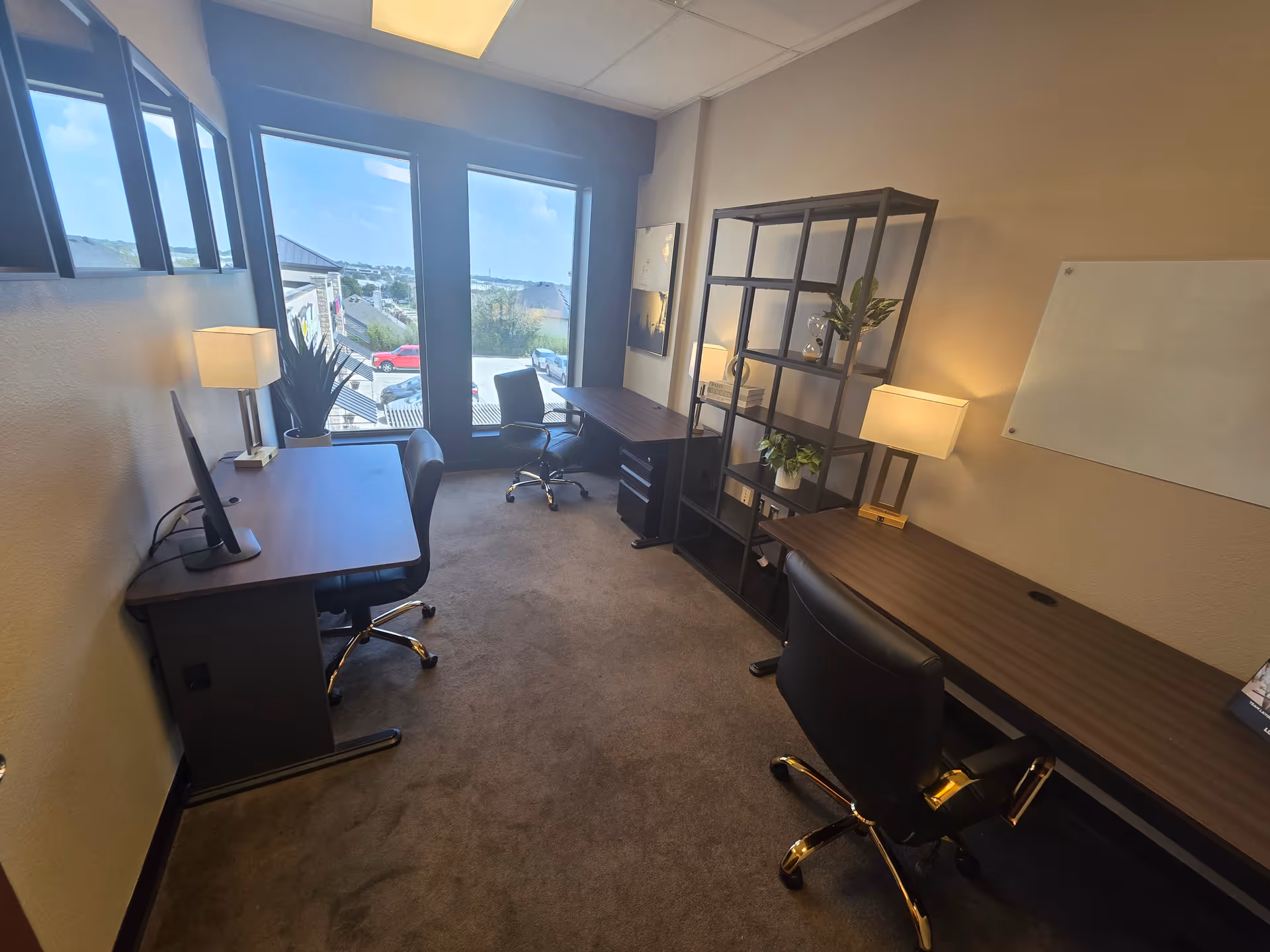 An interior office space with desks, chairs, shelving, plants, and a large window view.