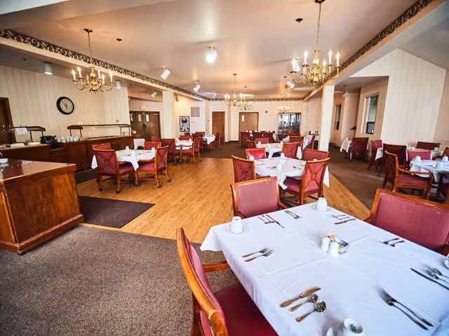 A spacious dining room with multiple tables covered in white tablecloths, each set with cups, utensils, and condiments. The room features wooden chairs with red cushions, chandeliers hanging from the ceiling, and a mix of carpet and wood flooring. There is a buffet area along one wall and a clock on the wall.