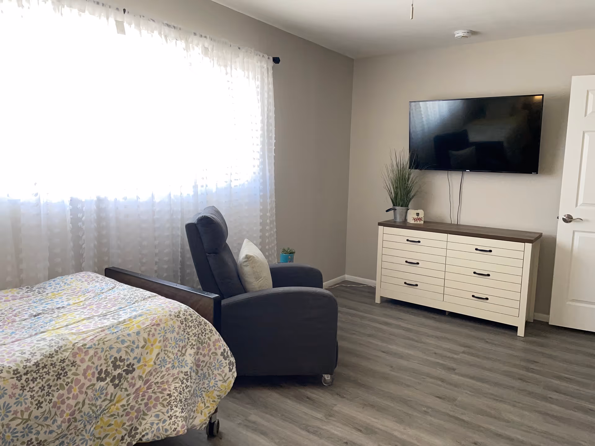 A furnished assisted living bedroom with a bed, gray recliner, dresser, and mounted TV.