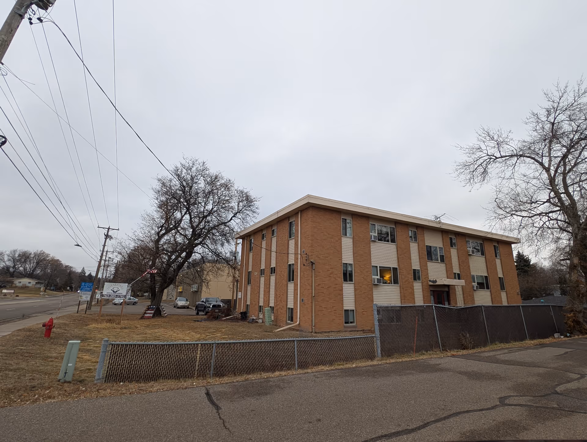 Exterior view of a three-story brick and beige panel building with multiple windows, surrounded by a chain-link fence and leafless trees on a cloudy day. A street with a fire hydrant and utility poles runs alongside the building.