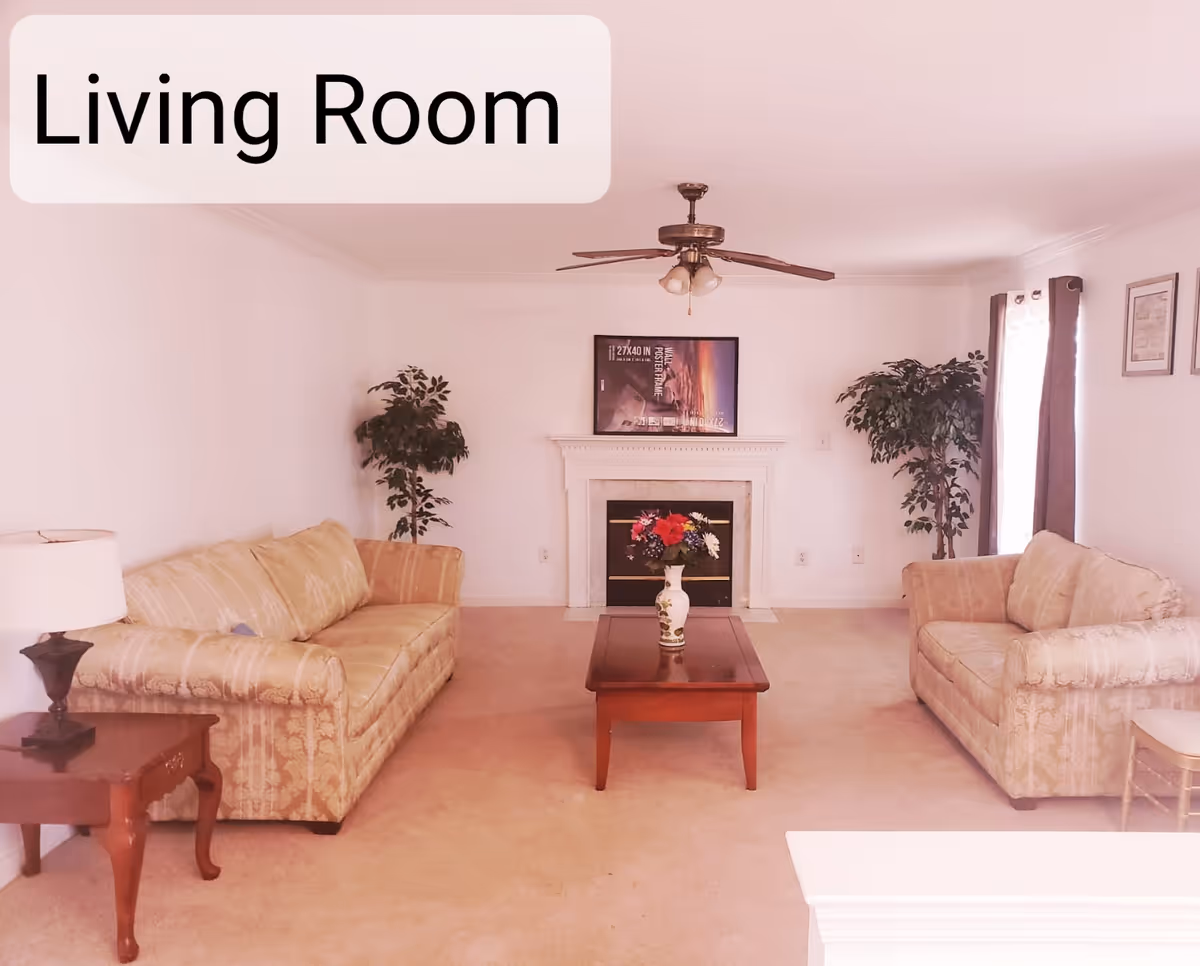 Cozy beige living room with two sofas, coffee table, fireplace, TV, ceiling fan, and plants.