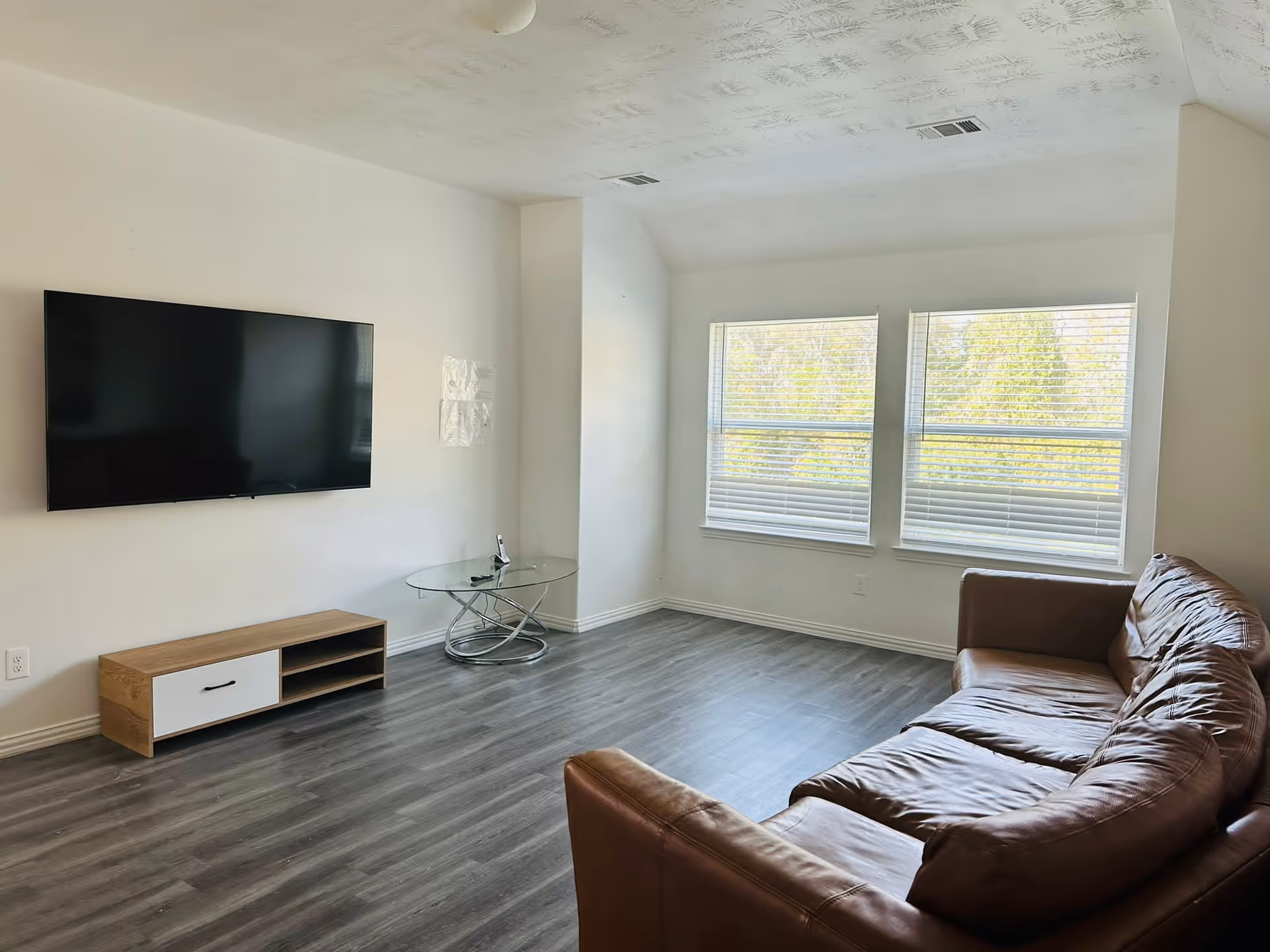 Modern living room with a wall-mounted TV, glass table, and brown leather sofa.