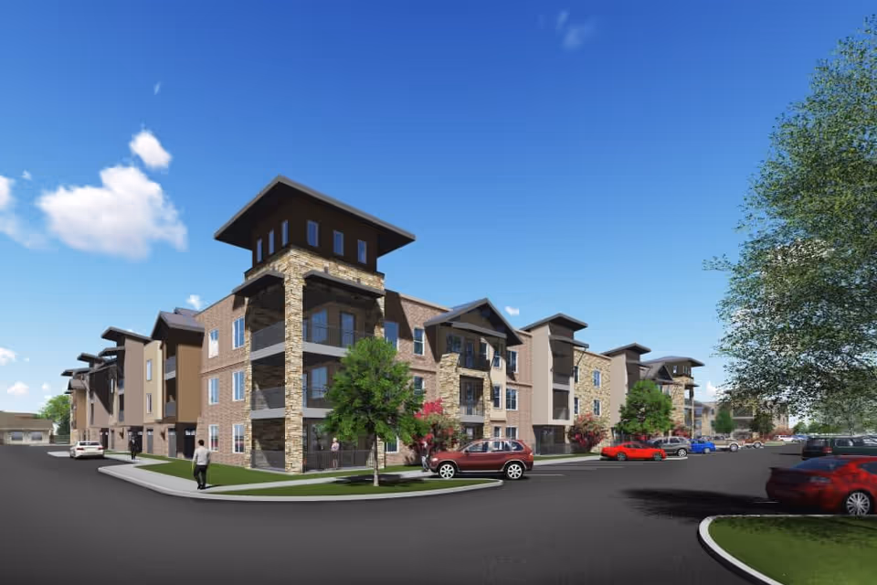 Exterior view of a modern multi-story senior living facility building with stone and brick facade, balconies, trees, parked cars, and a clear blue sky.