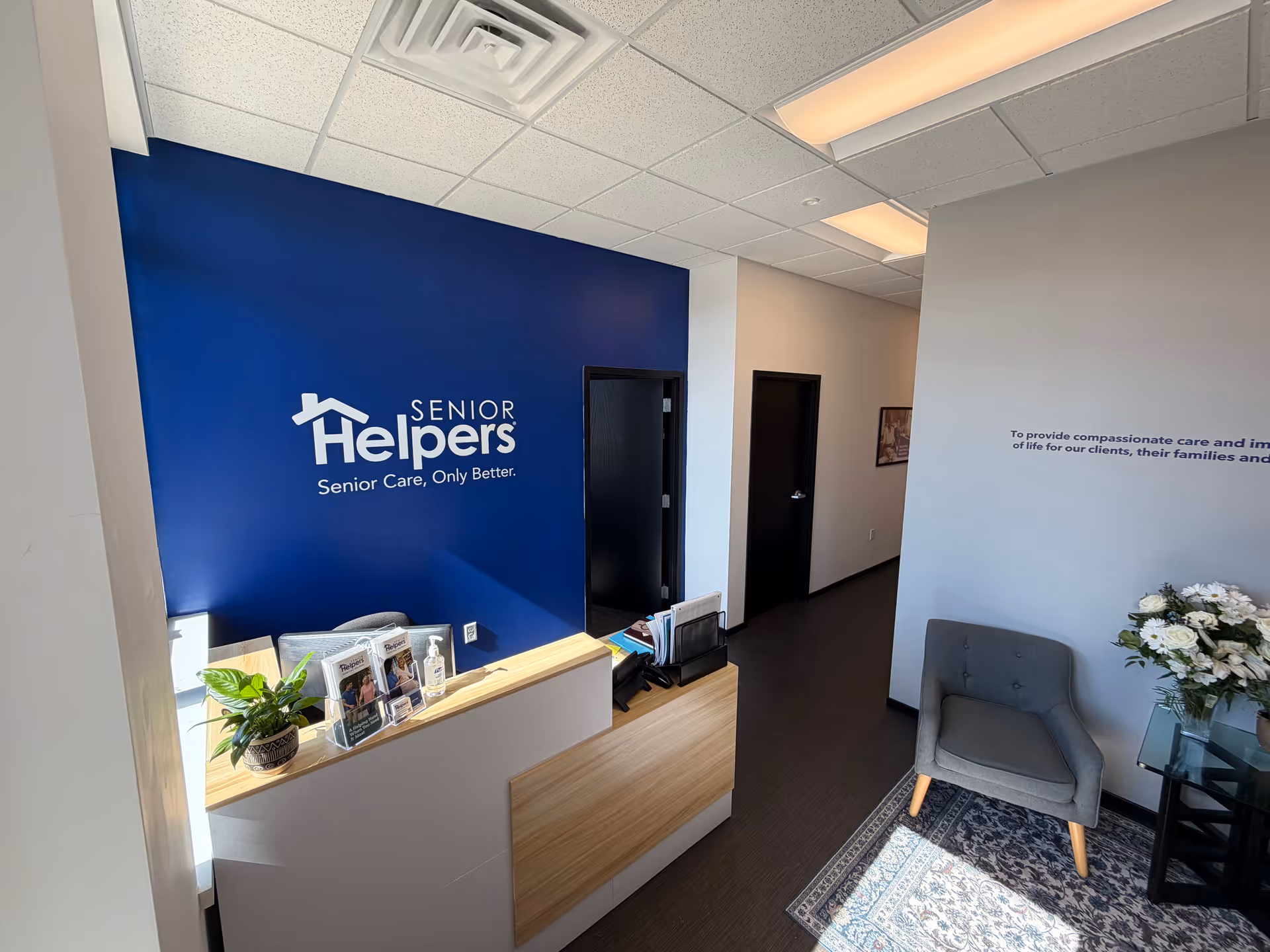 Reception lobby with blue wall and Senior Helpers logo, desk, plants, and seating area.