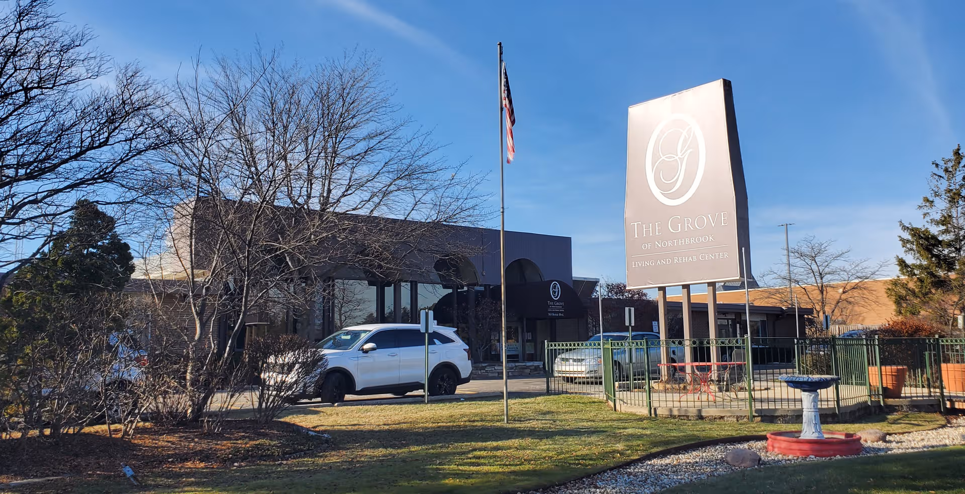 Front exterior of The Grove of Northbrook, with sign, flagpole, and parked cars.
