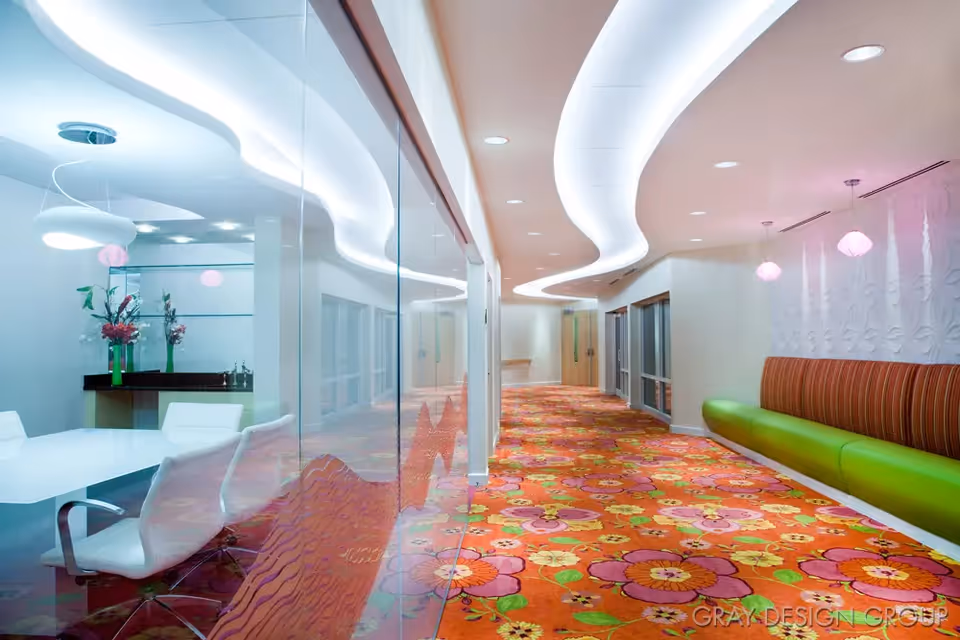 A brightly lit interior hallway with a colorful floral carpet. On the left side, there is a glass wall revealing a conference room with white chairs and a table, decorated with vases of flowers. On the right side, there is a long green bench with a striped backrest and hanging pendant lights above. The ceiling features a modern, wavy light design.