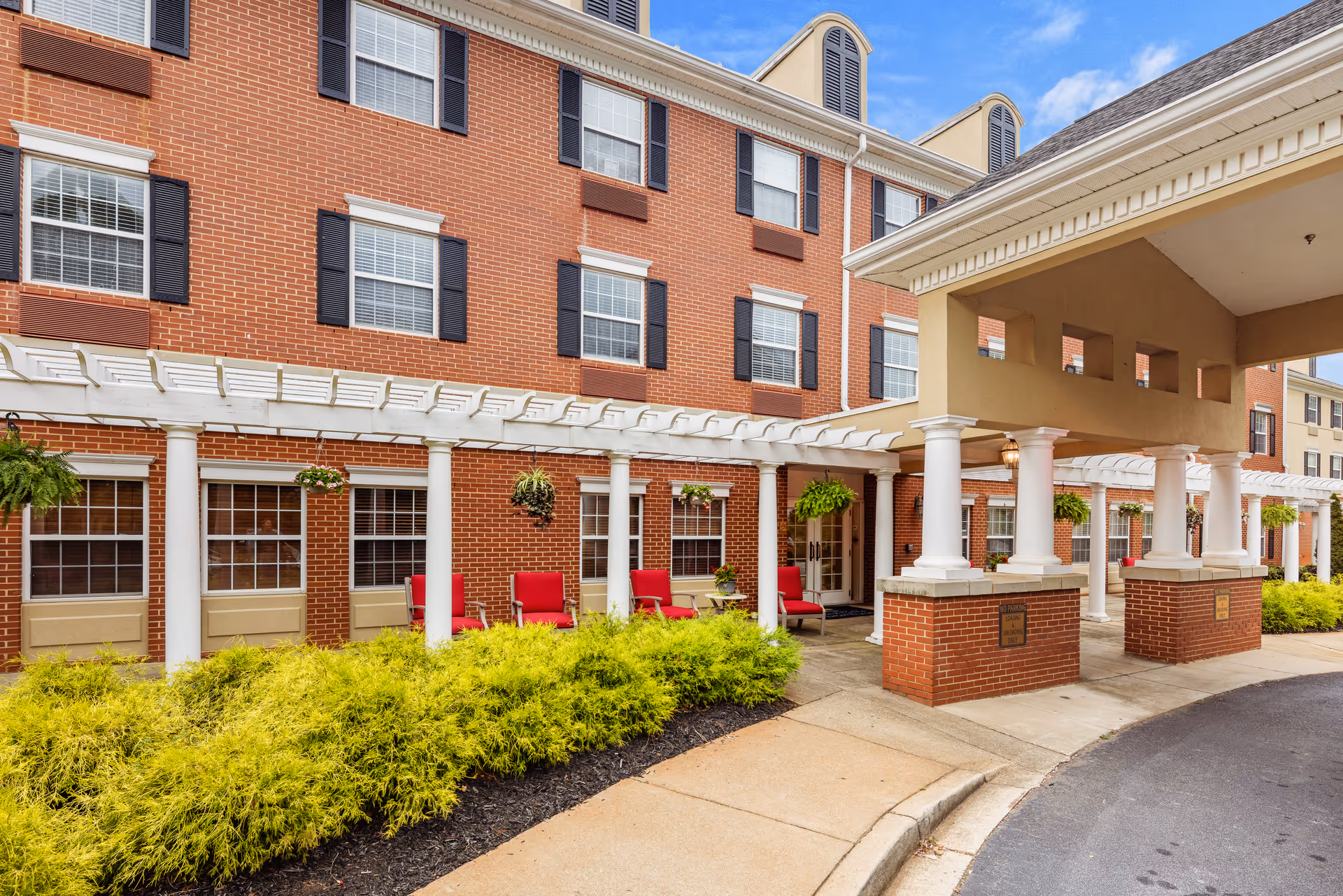 Exterior view of TerraBella Thornblade senior living facility showing a brick building with multiple windows, white columns supporting a covered entrance, a white pergola with hanging plants, red chairs arranged outside, and a landscaped area with green shrubs under a partly cloudy sky.