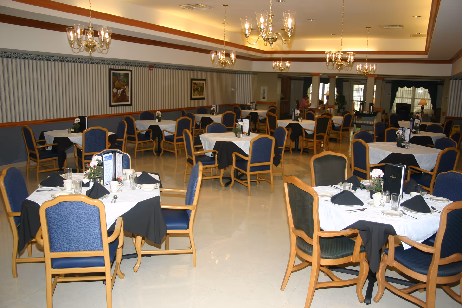 Large dining room with multiple tables set with white tablecloths, black napkins, wooden chairs, and chandeliers.