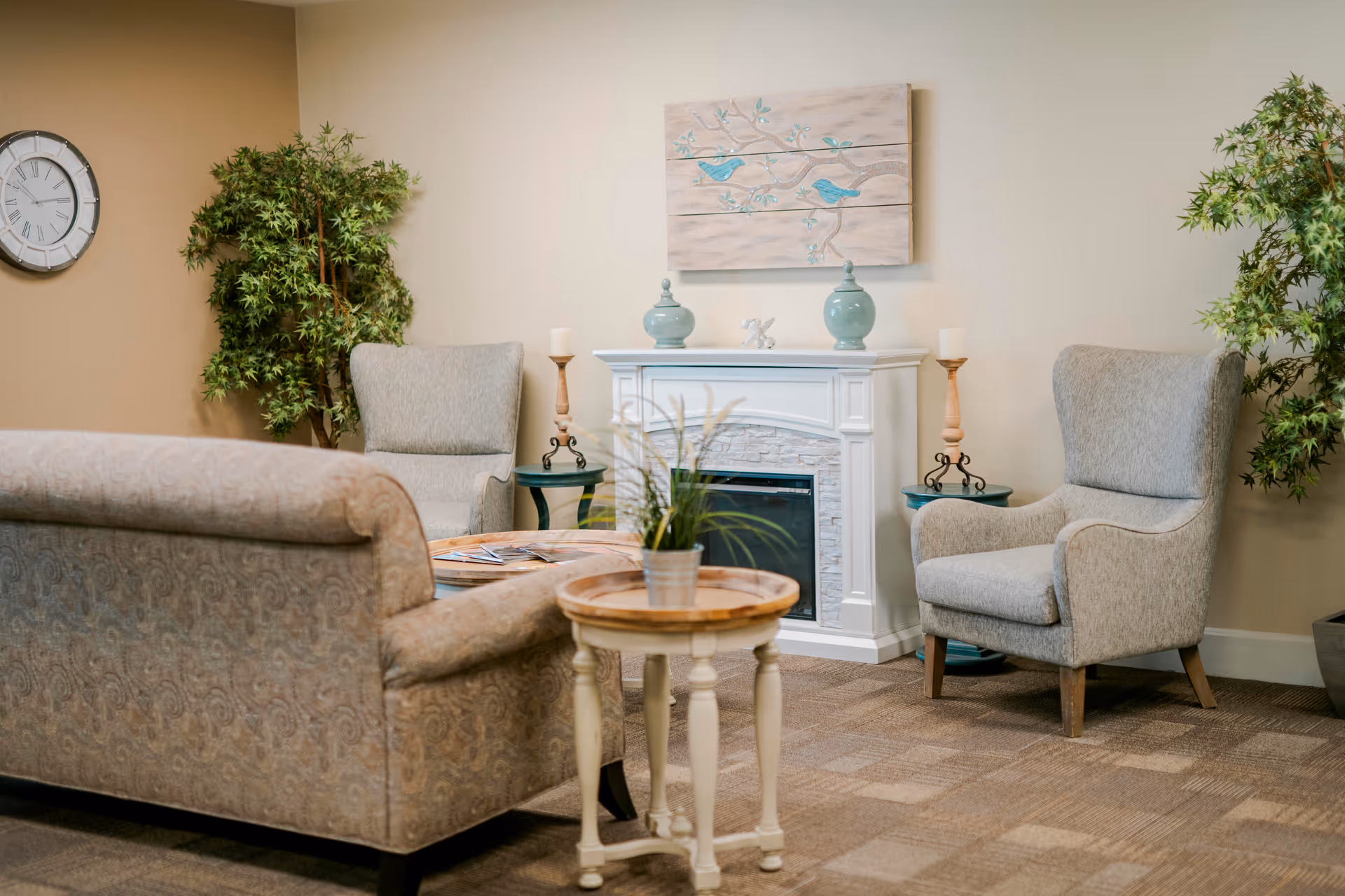 A cozy sitting area in a senior living facility with a patterned sofa, two gray armchairs, a white fireplace with decorative items on top, a round wooden side table with a potted plant, two small side tables with candles, a wall clock, and two large green potted plants against beige walls.