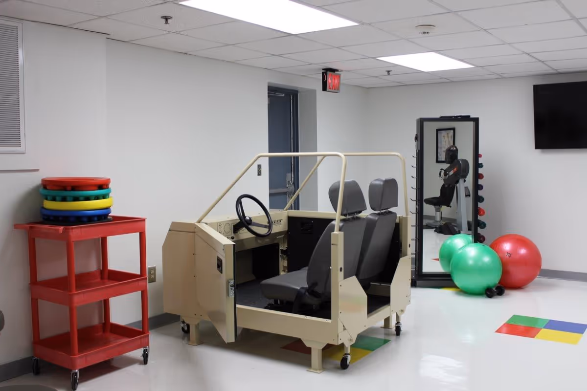 A therapy or rehabilitation room with a driving simulator setup featuring two seats and a steering wheel. Nearby are colorful balance cushions stacked on a red cart, two large exercise balls (one green and one red), a set of dumbbells, a full-length mirror, and an exercise machine. The room has a white ceiling with fluorescent lights and a white floor with colored square patterns.