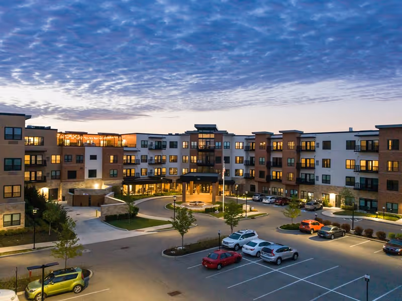 Exterior front view of a four-story senior living building at dusk with illuminated windows and parked cars in the lot.
