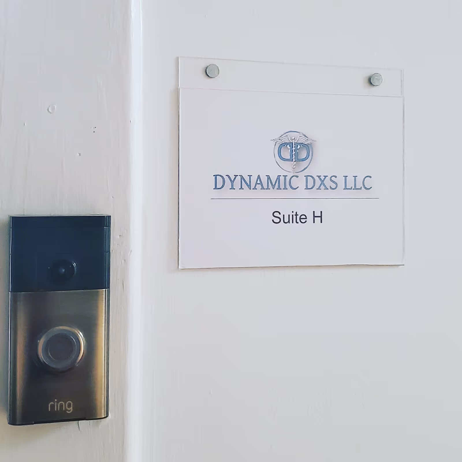Close-up wall scene with a sign reading Dynamic DXS LLC Suite H and a Ring doorbell.