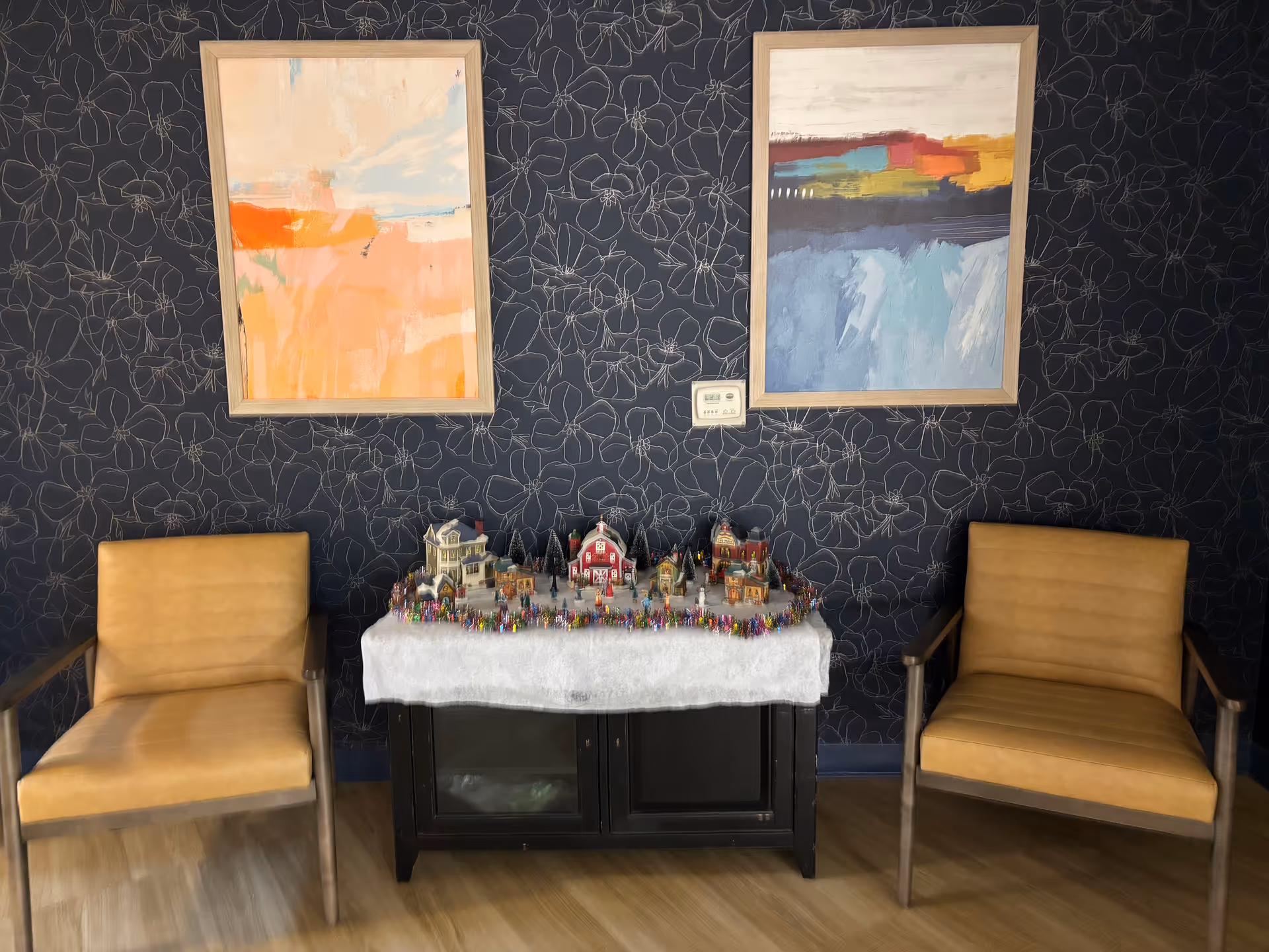 Two tan armchairs flank a table with a holiday village display against dark patterned wallpaper.