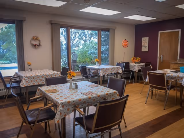 A dining room with several tables covered in patterned tablecloths featuring pumpkins and autumn leaves. Each table has chairs around it and small flower vases with yellow and orange flowers. Large windows show a view of trees and greenery outside. The room has wooden flooring and a purple accent wall with a door and wall decorations.