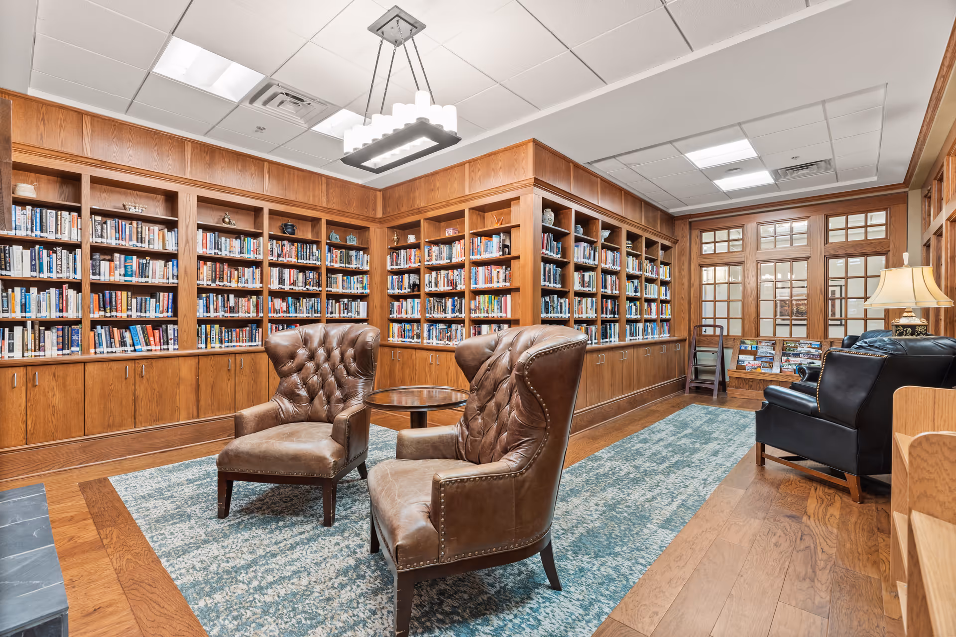 A cozy library room with wooden bookshelves filled with books lining the walls. Two brown leather tufted armchairs face each other with a small round wooden table between them. A blue patterned rug covers part of the wooden floor. In the background, there are large windows with wooden frames and a black leather armchair next to a table lamp.