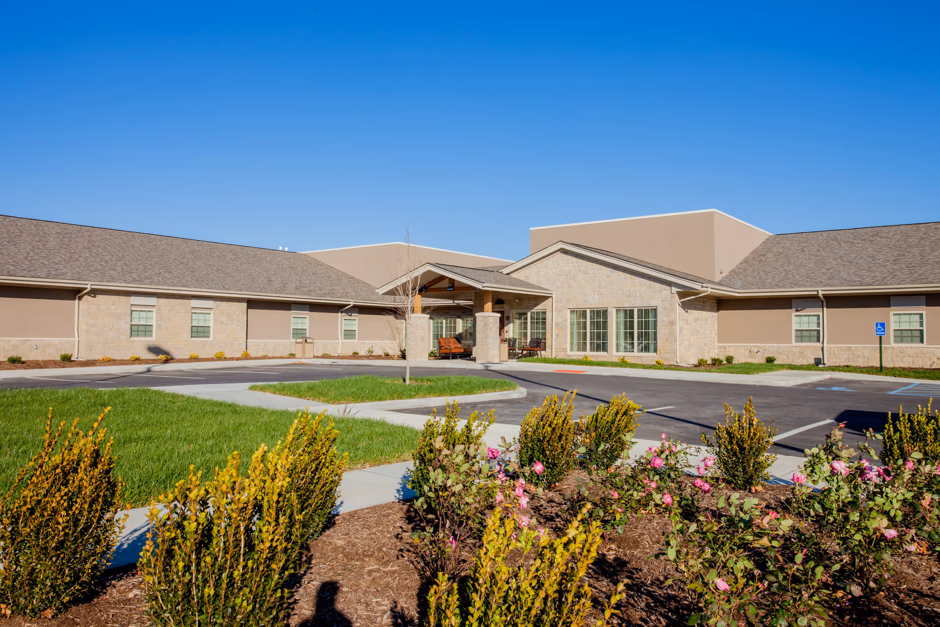 Exterior view of a single-story senior living facility building with a covered entrance, surrounded by a parking lot, green grass, and landscaped flower beds under a clear blue sky.