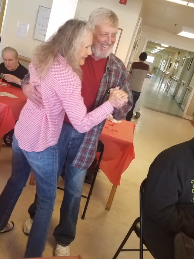 An elderly man and woman smiling and dancing together in a communal dining area of a senior living facility.