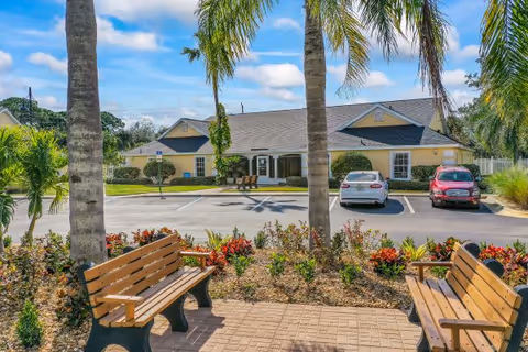 Exterior view of Hill Top Home of Comfort with benches, palm trees, and a parking lot.